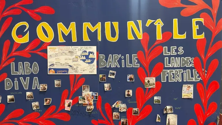 Un mur peint avec les couleurs du logo. Le fond est bleu, des plantes rouges serpentent le long du mur. En haut, Commun'île est écrit en gros, en jaune. Les différents noms de lieux sont écrits de part et d'autre, en gris. Des photos Polaroid de différents moments de vie sont collées sur le mur.