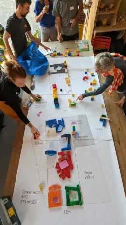 Une photo prise en plongée dans le Labo Diva. Le plan d'un futur bâtiment recouvre une grande table en bois. Des personnes sont debout autour, et participent à un atelier avec des Lego.