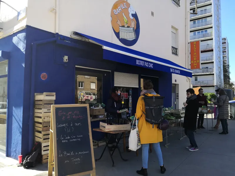 Le Bar Ile - Une terrasse d'un bar, les murs extérieurs sont bleus foncés. Un marché est installé avec des légumes.